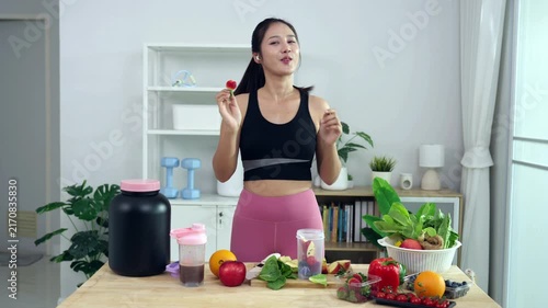 Smiling Asian fitness influencer recording a health podcast, scooping pink protein powder into a shaker bottle. Woman vlogger creating sports nutrition and healthy diet content with fresh food.