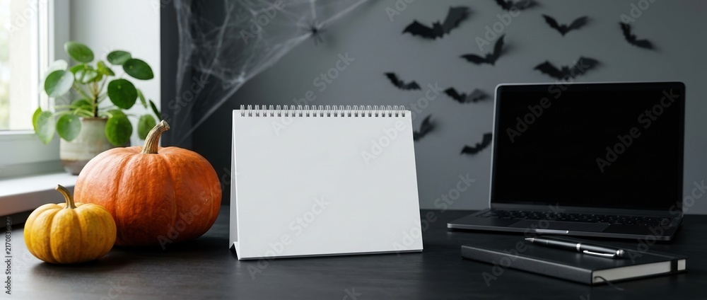 horizontal close-up of Halloween-themed workspace with blank calendar, notebook, desk, laptop and pumpkin. modern, tidy home office environment with a thematic Halloween spirit
