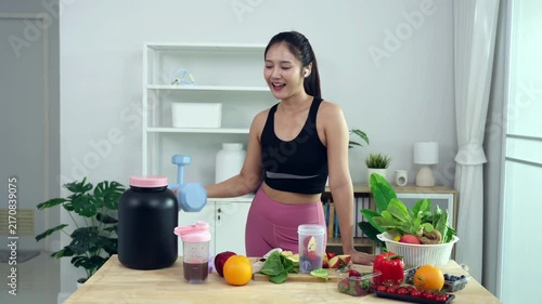 Smiling Asian fitness influencer recording a health podcast, scooping pink protein powder into a shaker bottle. Woman vlogger creating sports nutrition and healthy diet content with fresh food.