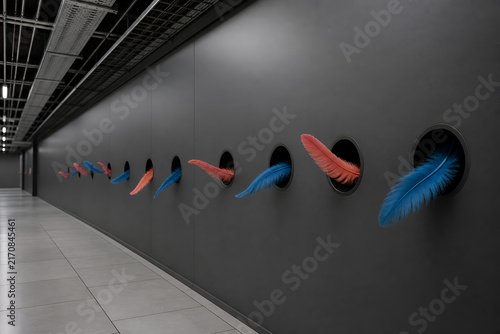 Playful feathers emerge from circular holes in a hallway with dark walls and tile flooring, reflecting a lighthearted approach to technology and design