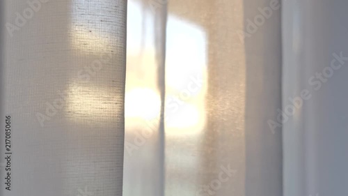 Handheld camera moving past sheer white curtains glowing in warm evening sunlight as the fabric gently sways in a summer breeze.