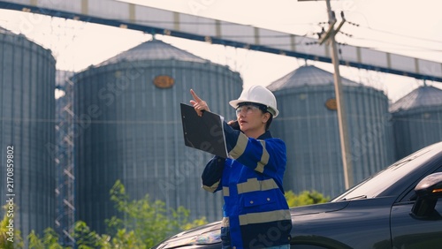Asian female engineer in safety helmet inspecting industrial grain storage silo plant with laptop. Agribusiness engineering audit, factory facility management, ESG compliance and logistics concept.