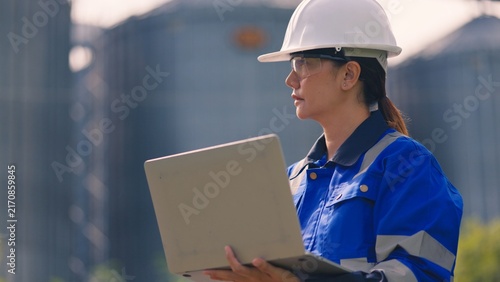 Plakat Asian female engineer in safety helmet inspecting industrial grain storage silo plant with laptop. Agribusiness engineering audit, factory facility management, ESG compliance and logistics concept.