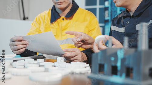 Two Asian male industrial engineers, wearing helmets, safety glasses, and uniforms, are working together in a lab. They are inspecting and analyzing white mechanical gear parts printed with a 3D print