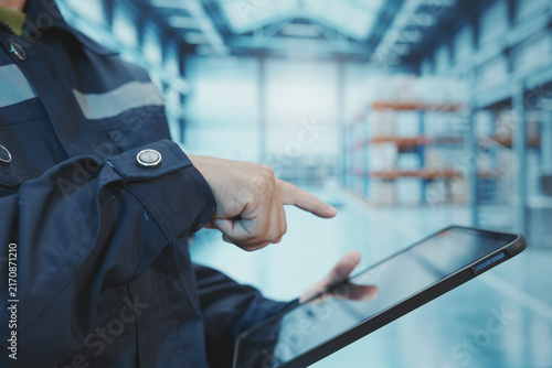 A close-up shot shows a factory worker using their finger to control a digital tablet. The backdrop is a large, modern manufacturing plant with automated production lines, shelving, control systems, a