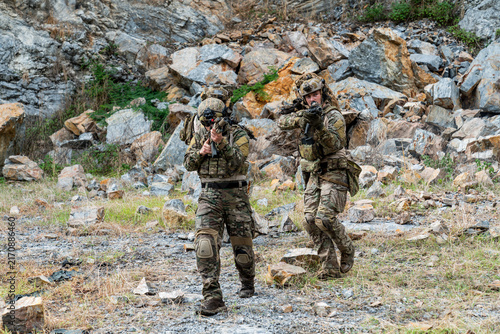 Military personnel engage in tactical training in a rocky outdoor area during daylight
