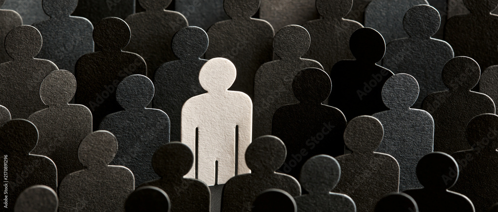 Fototapeta premium A prominent white human silhouette stands distinctly within a vast and textured crowd of dark gray and black abstract paper cutouts, powerfully symbolizing individuality, uniqueness, and