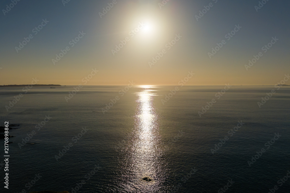 Obraz premium Golden evening sunlight reflecting across calm ocean surface at sunset, gentle swell on sea with distant headland on horizon under clear dusk sky