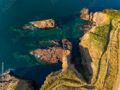 Rocky cliffs and red sandstone sea stacks on Pembrokeshire coastline, Wales, warm evening sunlight illuminating grassy headland and turquoise water, aerial view