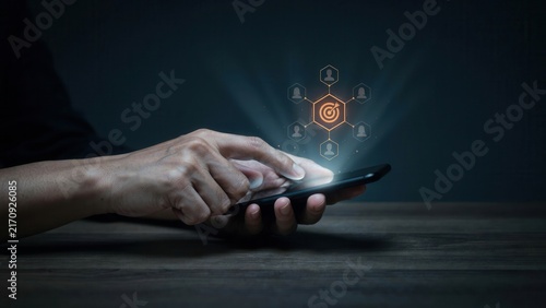 Business professional interacting with smartphone screen showing holographic target marketing icons representing customer engagement strategy in dark office lighting environment setting