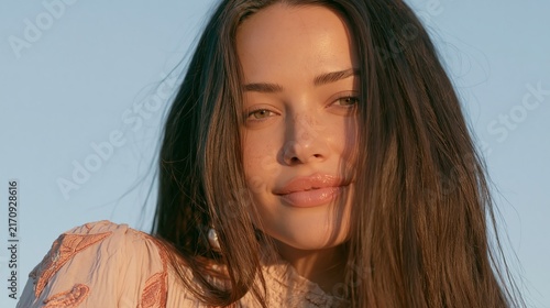 Close up portrait of a beautiful young woman with long brown hair smiling softly against a clear blue sky background during golden hour lighting.