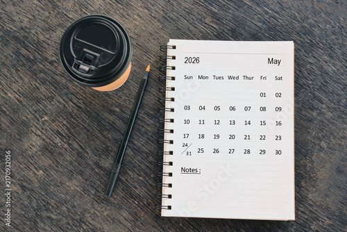 May 2026 desk calendar with disposable coffee cup and pen on wooden table. Directly above. Flat lay.