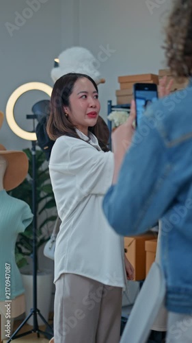 Young Asian woman fashion entrepreneur live streaming selling clothes online with smartphone ring light and laptop in home studio, representing small business owner and professional women.