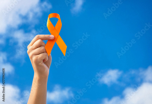 A hand holding an orange awareness ribbon up against a clear bright blue sky representing solidarity and support for important social causes