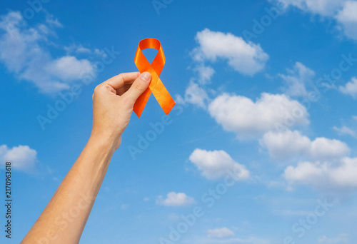 A hand holding an orange awareness ribbon isolated against a bright blue sky with soft white clouds representing support and hope