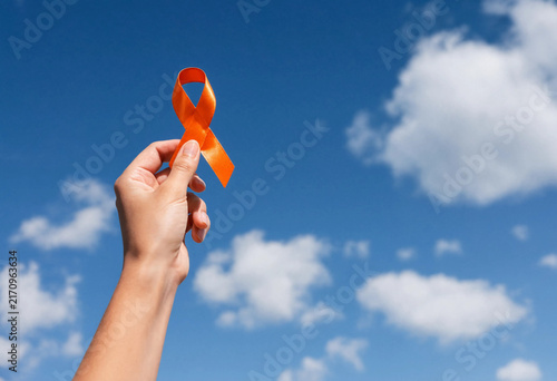 A hand holding up a vibrant orange awareness ribbon against a beautiful blue sky with soft white clouds symbolizing support and solidarity