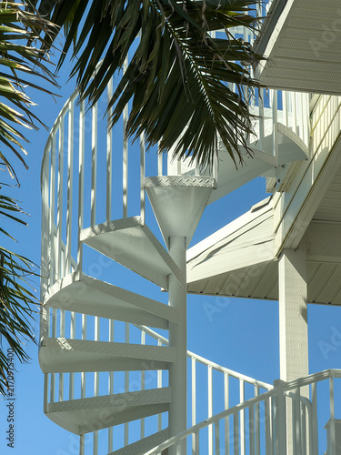 An exterior spiral staircase, shaded by palm trees, leads to an observation deck on the roof of a beach house with a seaside view on a barrier island in Florida. Example of architectural escapism.