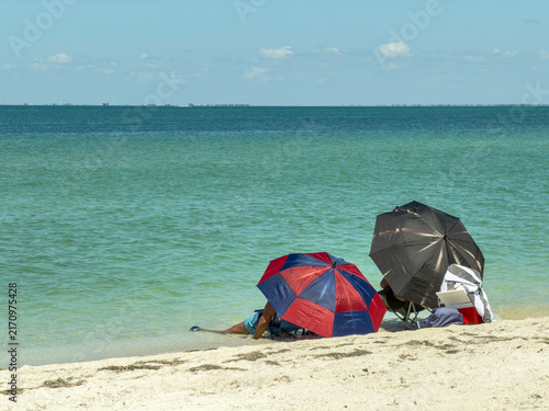 Two dark beach umbrellas shade two adult beachgoers relaxing by Sarasota Bay on a bright, sunny afternoon in summer along the Gulf Coast of Florida