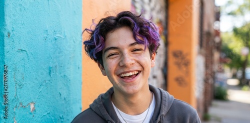 Portrait of happy teenage boy with purple curly hair laughing outdoors against graffiti wall. Joyful youth expression, positive emotion and generation z lifestyle concept