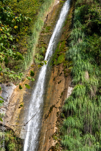 Long white waterfall cascading down a moss-covered rocky slope surrounded by lush green vegetation.