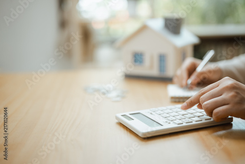 Photography A person uses a calculator beside a model house while writing figures to estimate mortgage costs and compare each loan option before choosing an affordable monthly amount for a planned purchase
