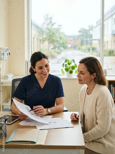 Woman in blue scrubs gesturing with a pen at a wooden office desk indoors