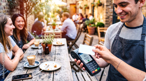 Customer paying contactless bill with credit card on POS terminal held by smiling waiter in outdoor cafe patio