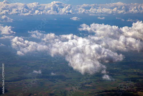 An aerial view of a cloudscape,  scattered puffy cumulus or stratocumulus clouds hovering above a patchy, green landscape visible on the ground below. Mid-to-low level detached cumulus clouds. 