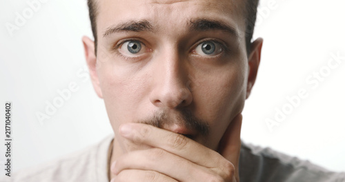 Quadro su tela Portrait of a shocked young Caucasian man with a mustache covering his mouth against a white studio background