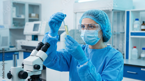 Laboratory female scientist in protective gear inspecting a liquid sample in a test tube beside microscope during biomedical research and diagnostic analysis.
