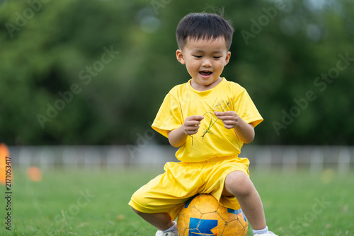 Joyful Asian child sitting on football during daytime sports practice on green grass field, authentic playful toddler activity