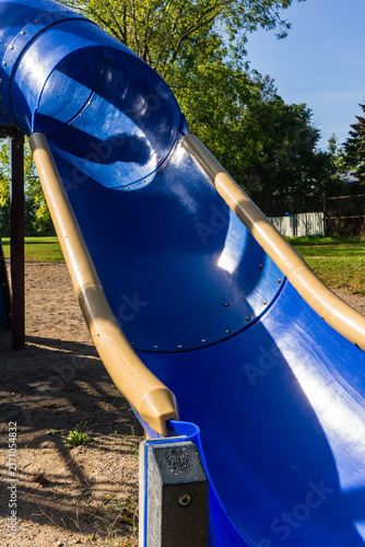 Bright blue spiral tube slide in a sunny outdoor playground surrounded by trees and greenery