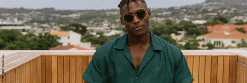 Fototapeta premium Young man in green shirt and sunglasses on rooftop terrace for lifestyle banner and social media background with urban view