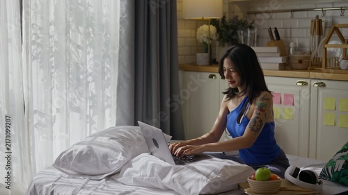 Freelancer woman Gen Z sitting on bed and typing on a laptop in apartment.asian female remote work in kitchen at home.digital nomad,online work,remote working and video conference training concept
