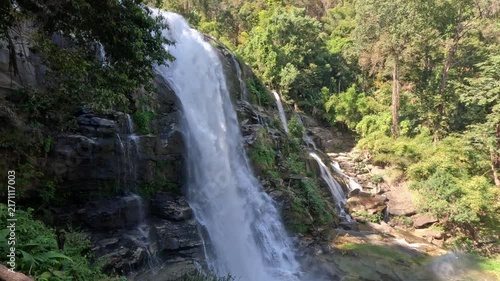 A wide shot of a powerful waterfall flowing rapidly over rocks in a protected natural area in northern Thailand, surrounded by dense tropical forest and lush greenery.