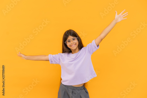 happy little brunette girl in glasses is having fun, raising her hands up against a yellow background in a studio, with space for text