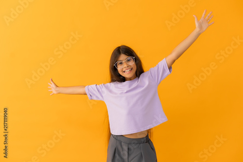 happy little brunette girl in glasses is having fun, raising her hands up against a yellow background in a studio, with space for text