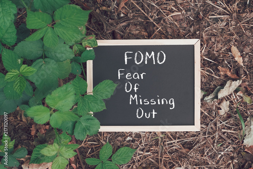 Chalkboard displaying the message 'FOMO Fear Of Missing Out' placed on a forest floor surrounded by green leaves and dry foliage, suggesting the concept of social anxiety and nature.