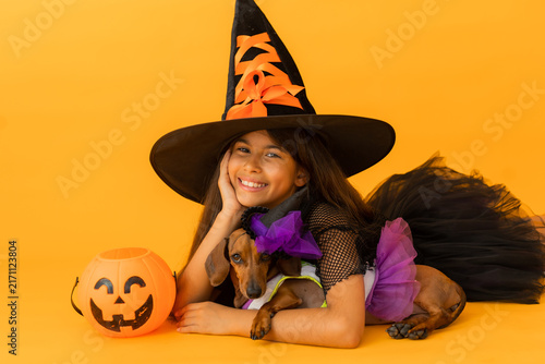 little brunette girl in a witch costume for Halloween against a yellow background in a studio with a dachshund dog, space for text.