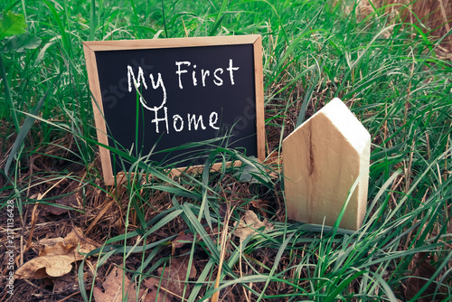 A small chalkboard sign with the words 'My First Home' placed in lush green grass next to a wooden house shaped figure, symbolizing homeownership or real estate investment.