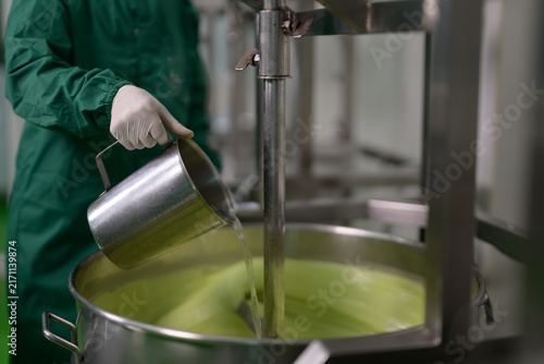 A person in protective gear pours liquid into a large stainless steel container, likely in a controlled industrial or laboratory setting.