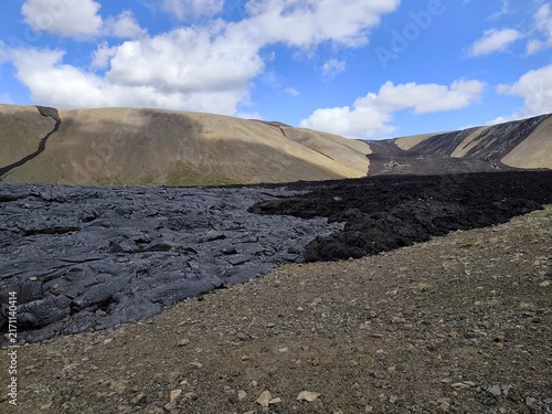 Картината върху платно Black solidified lava of a recent eruption fills a valley below eroded volcanic hills under a blue sky with clouds, Fagradalsfjall area, Iceland