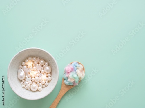 Minimalist surreal flat lay: ceramic bowl filled with glowing pearls and wooden spoon holding pastel rainbow cotton on mint background