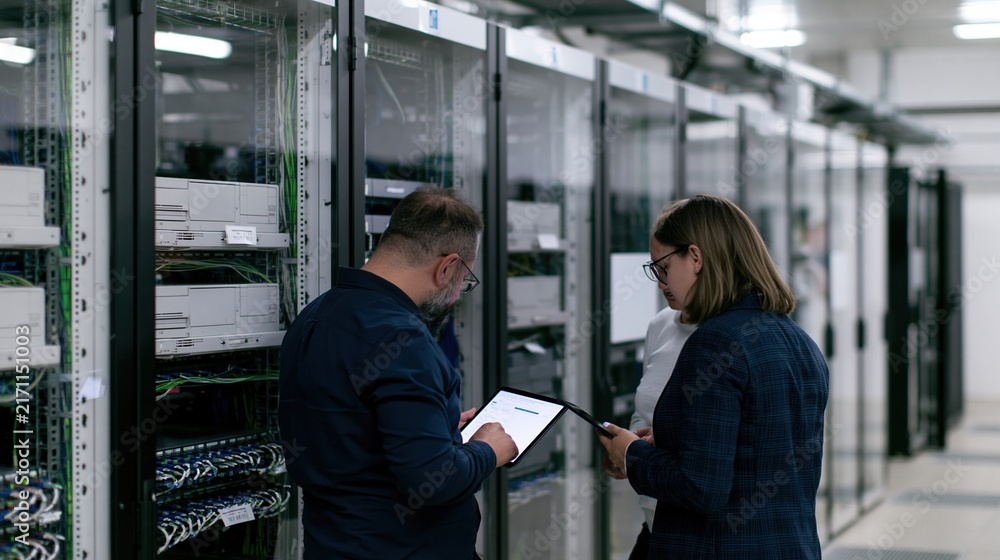 Fototapeta premium Server room with IT professionals discussing data on a tablet, featuring glowing panels and brushed steel surfaces for cybersecurity and maintenance