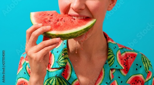 Woman eating fresh juicy watermelon slice wearing patterned summer shirt against vibrant bright blue background with tropical