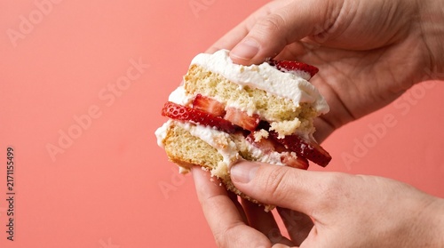 Hands holding fresh strawberry shortcake with whipped cream against pink background with person holding piece indulgence