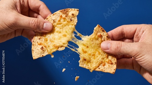 Hands pulling apart warm crispy tortilla toasted with melted gooey stretchy cheese against vibrant blue background with snack