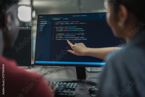 Male and female developers discuss programming solution together, pointing at lines of code on monitor while working as small software team in dim office