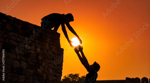 Silhouette of man helping another climb over stone wall during sunset with person assisting another dark silhouette one