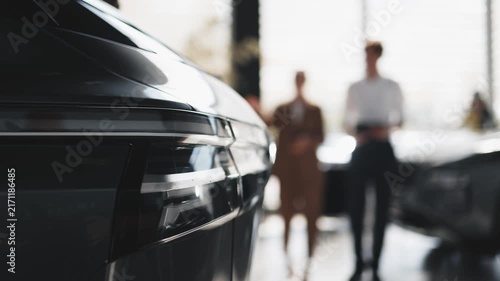 Front detail of a new car in modern dealership while salesperson talks with female customer interested in purchasing a vehicle